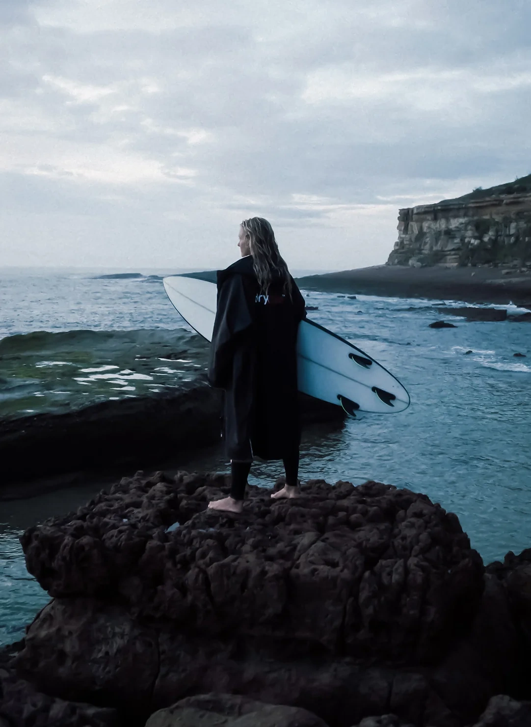 Adult surfer looking out to sea with surfboard, wearing long sleeve Dryrobe® Advance