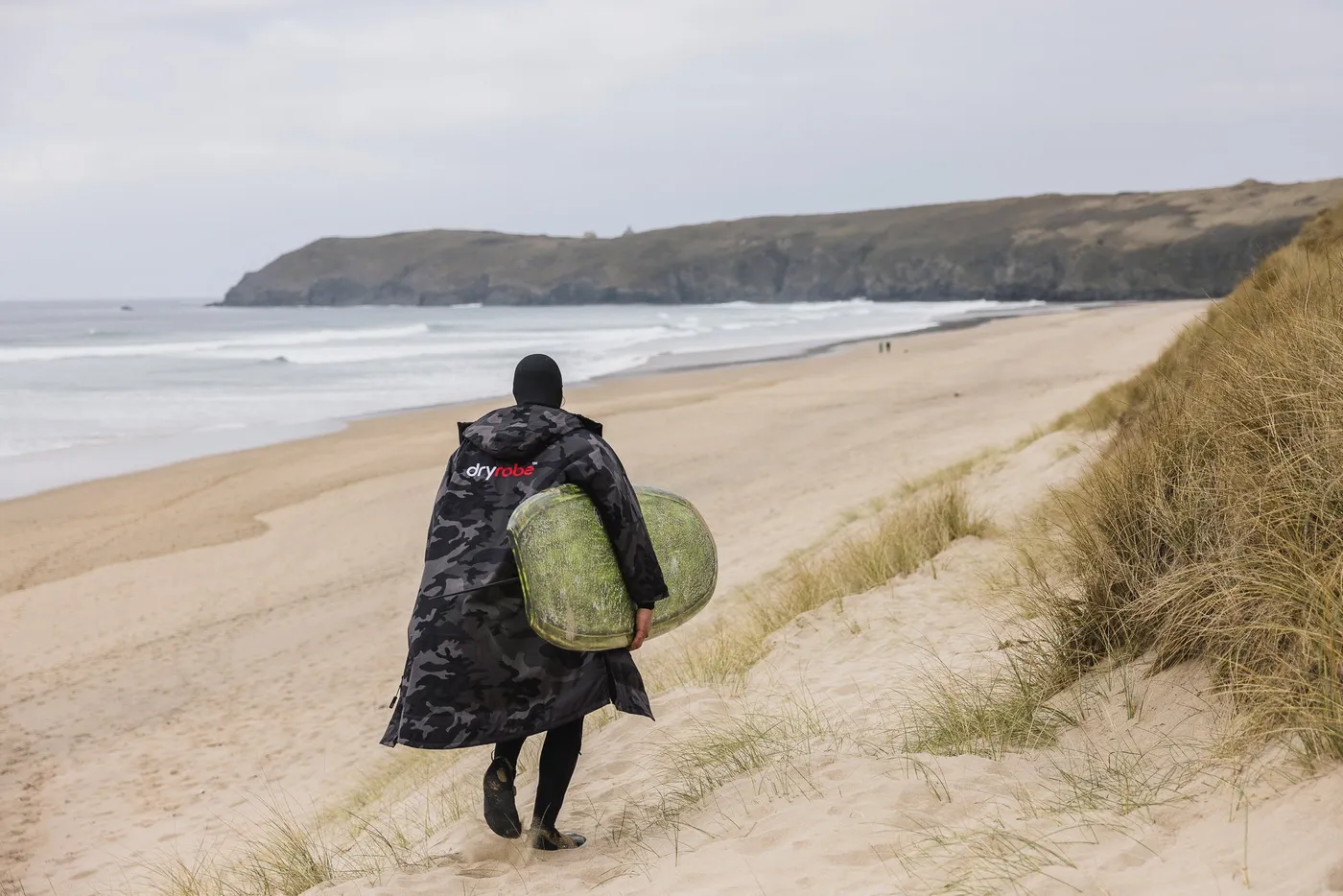 A surfer walking down the beach in a wetsuit and Dryrobe carrying a surfboard