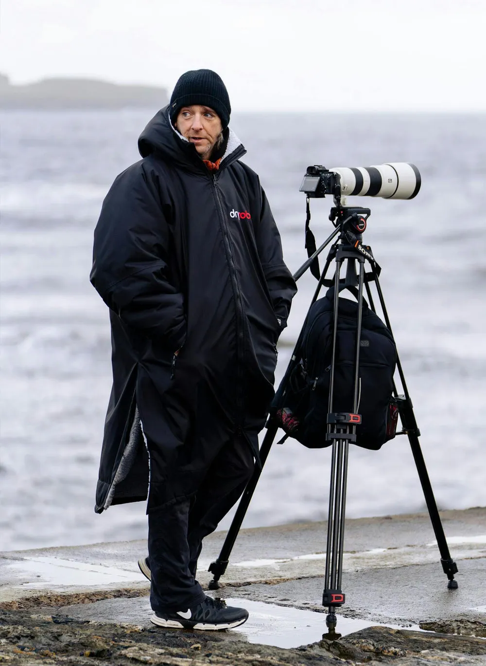Person in a long black Dryrobe standing beside a camera on a tripod at the coast