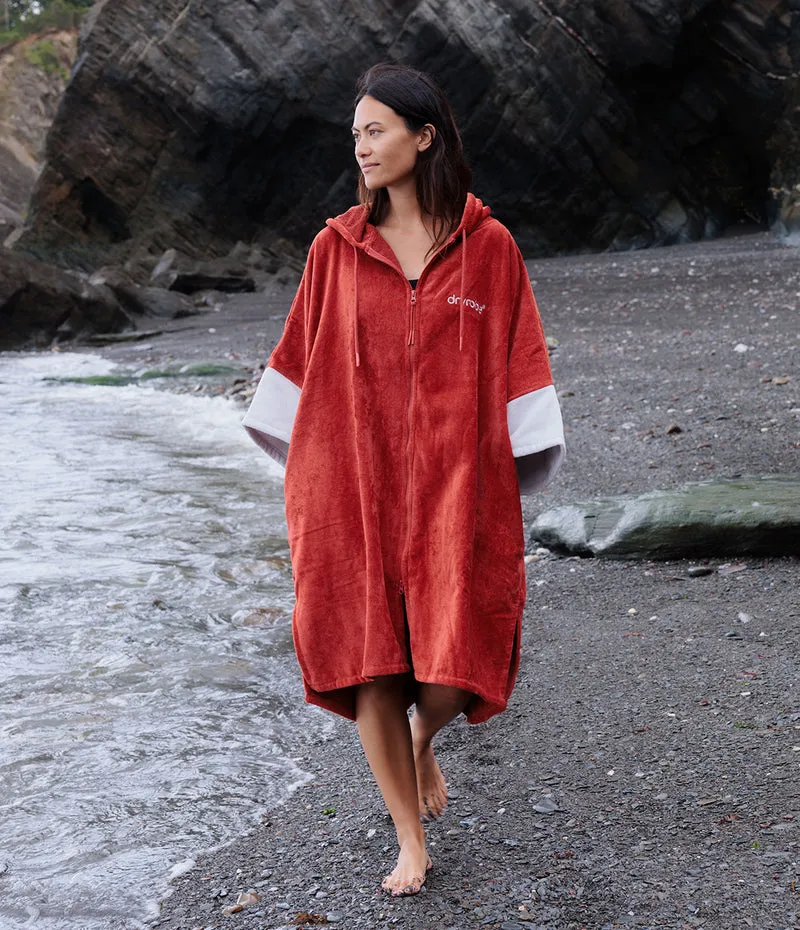 A woman walking barefoot on a rocky beach wearing a terracotta red hooded towel poncho with white sleeve accents.