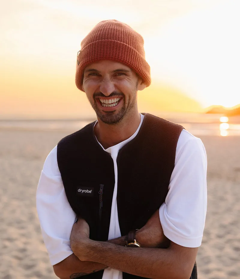 A smiling man on a beach at sunset wearing a rust-orange knit beanie, a white t-shirt, and a black Dryobe fleece vest.