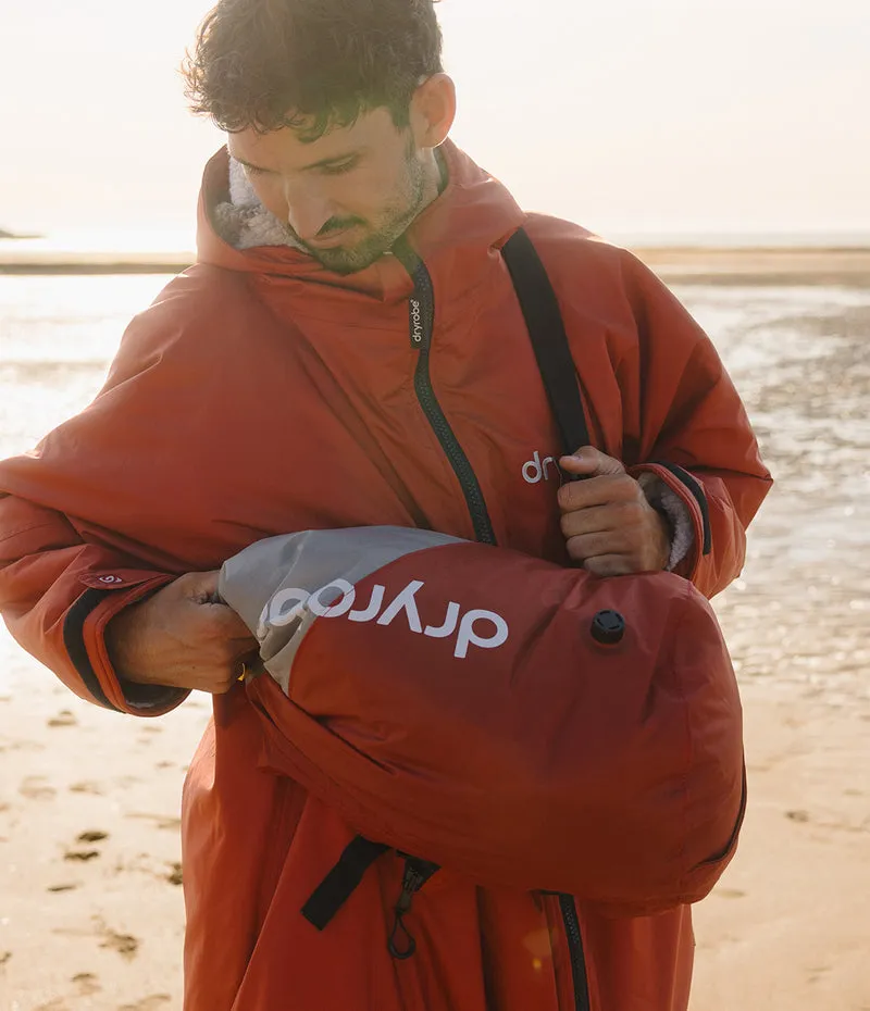 A man on a sandy beach wearing a rust-orange waterproof suit and holding a gray Dryrobe dry bag pouch.